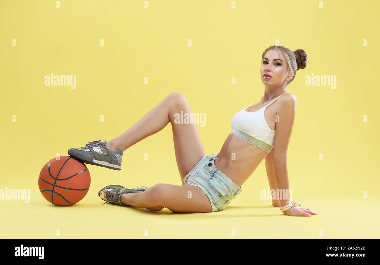 Montare Pretty girl indossano jeans corti seduto sul pavimento con la pallacanestro su sfondo giallo Foto Stock