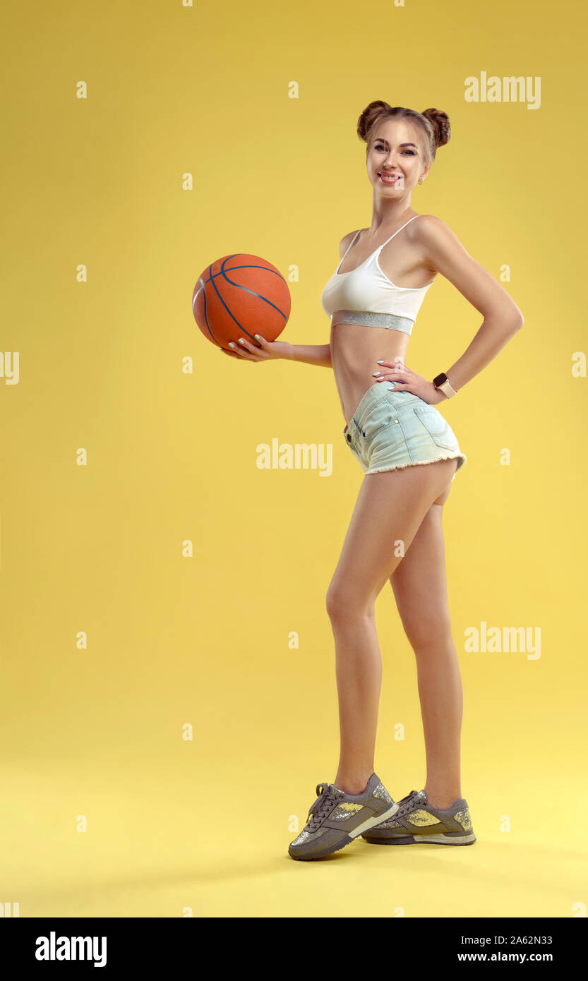 Ragazza che indossa jeans corti tenendo una palla da basket e posa su sfondo giallo Foto Stock