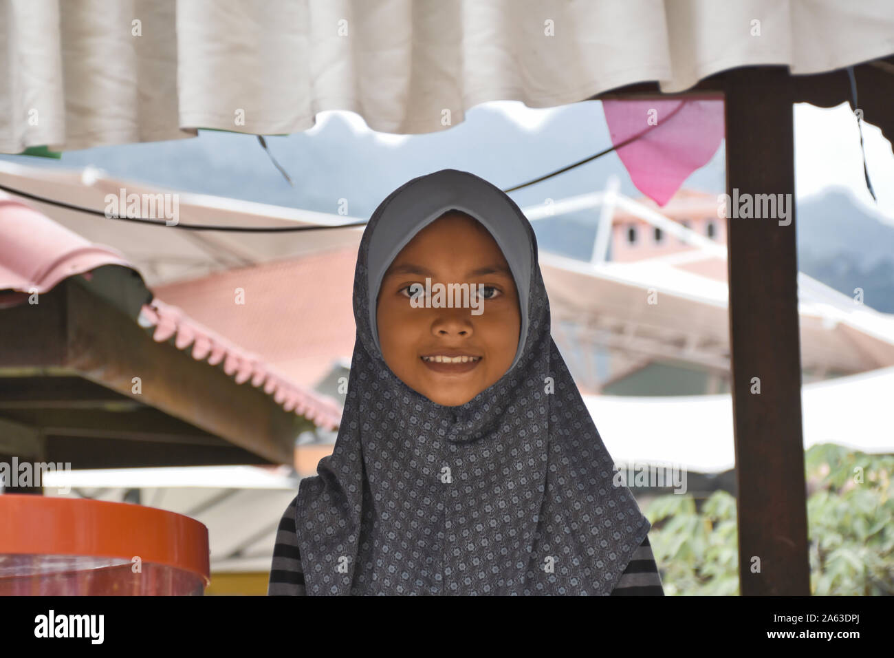 Il Langkawi, Malesia 08.12.2019: una bella e gentile e innocente malese giovane ragazzina musulmana in tradizionale hijab sorridente e aiutare la sua madre la vendita Foto Stock
