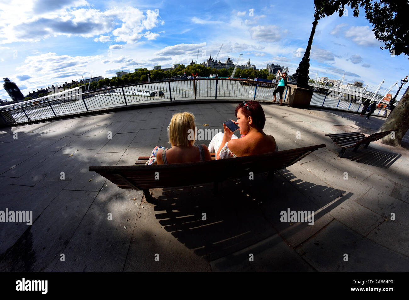Londra, Inghilterra, Regno Unito. Due giovani donne seduta su una panchina sulla riva sud, guardando oltre il Fiume Tamigi Foto Stock