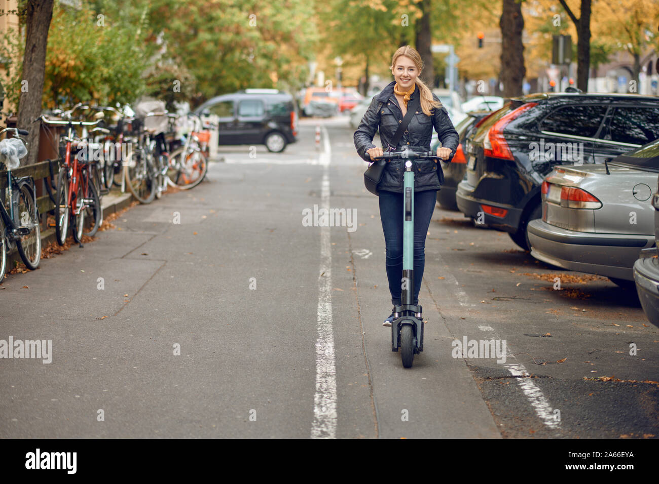Felice giovane donna bionda in sella ad una scooter elettrico nella città, sorridente alla fotocamera, in autunno Foto Stock