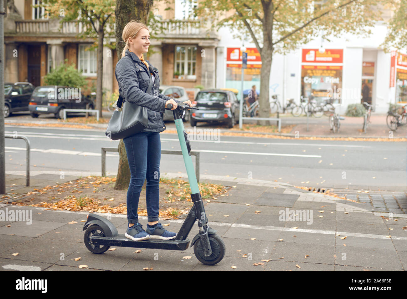 Felice giovane donna bionda in sella ad una scooter elettrico in città in autunno, vista laterale Foto Stock
