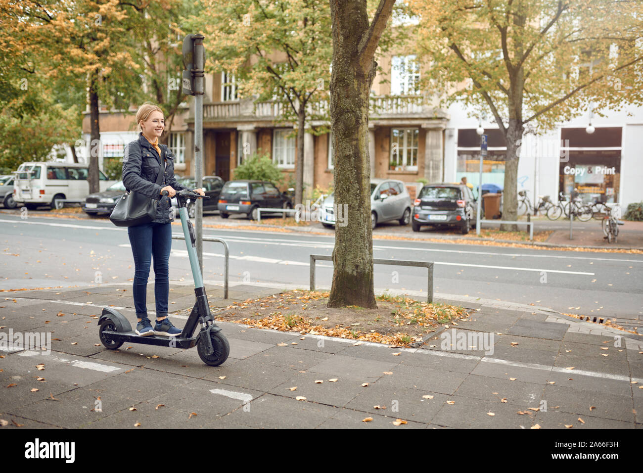 Felice giovane donna bionda in sella ad una scooter elettrico in città in autunno, vista laterale Foto Stock