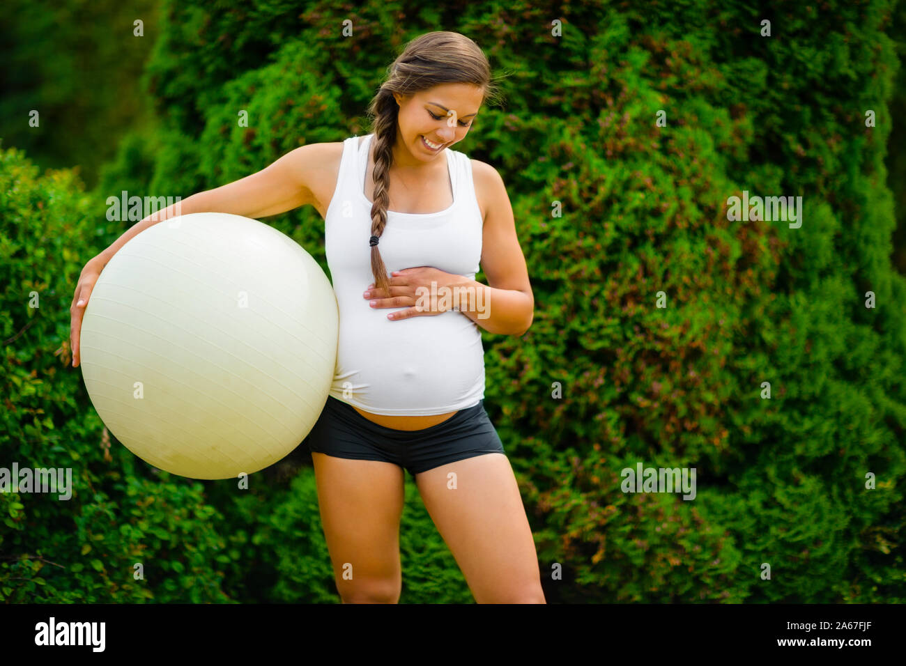 La mamma che si attende tocca lo stomaco mentre tiene la palla da ginnastica nel parco Foto Stock