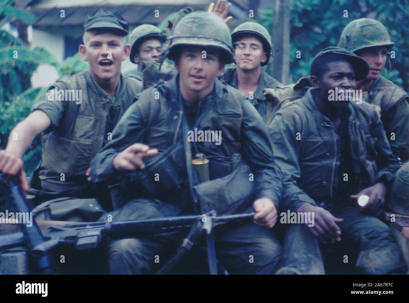 Un gruppo di soldati americani armati e uniformati sorridono, ondono e puntano verso la telecamera, probabilmente mentre passano sul letto di un veicolo di trasporto militare, Vietnam, 1965. () Foto Stock