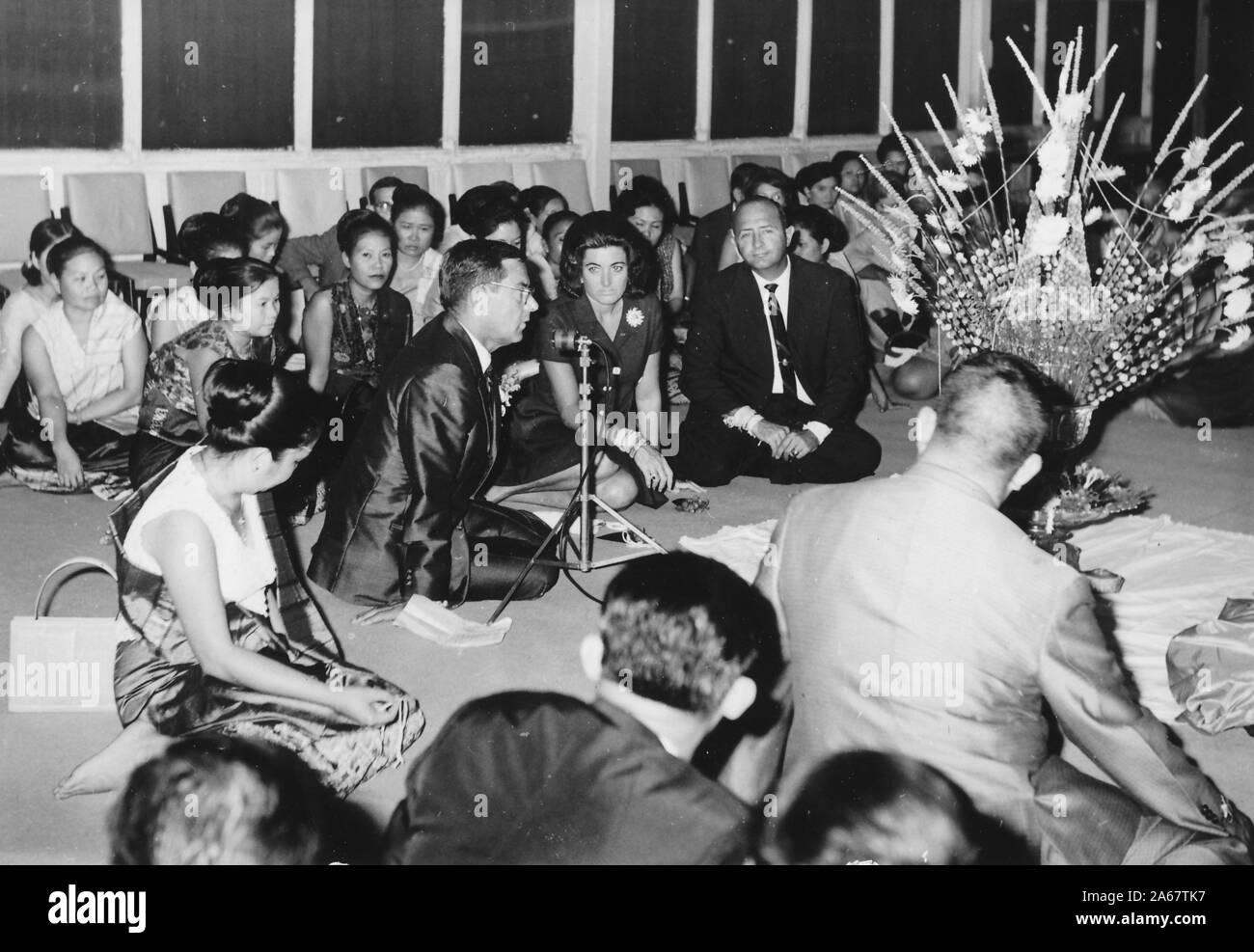 Un gruppo di uomini e donne formalmente vestiti siedono sul pavimento ascoltando un altoparlante durante un evento condiviso per l'esercito americano e l'esercito tailandese reale, fotografato durante la guerra del Vietnam, 1968. () Foto Stock