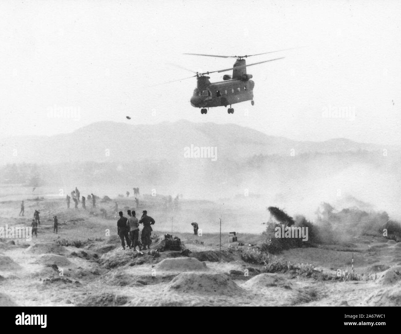 Un elicottero Boeing CH-47 Chinook solleva la polvere mentre vola sopra una zona leggermente collinare circondata da soldati che guardano e camminano lontano dalla nube di polvere, Vietnam, 1965. () Foto Stock