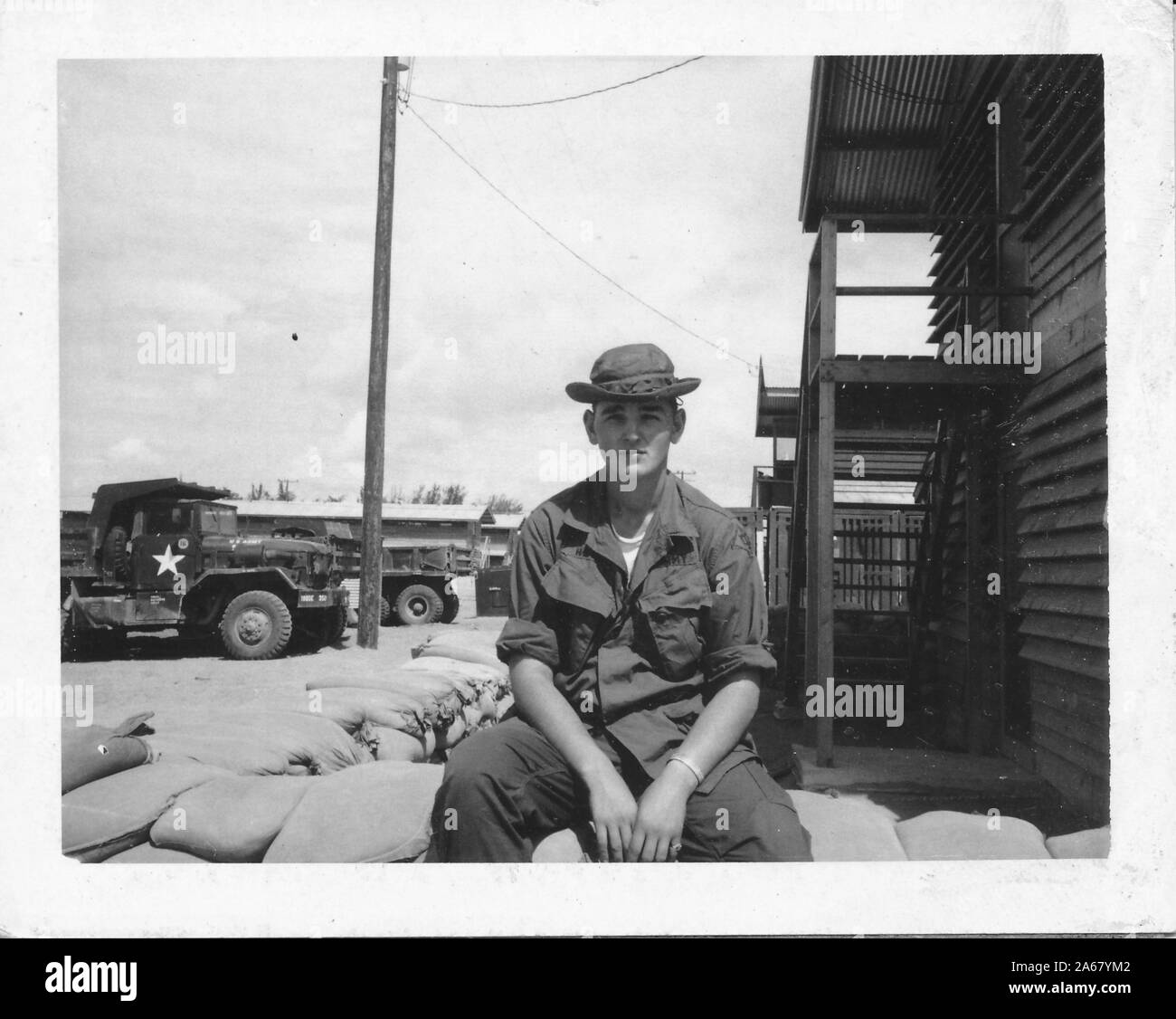 A metà colpo di Army SPC Gary Heizer, seduto fuori su una parete bassa di sacchi di sabbia in un campo militare, di fronte alla macchina fotografica con un'espressione rassegnata sul suo volto, Vietnam, 1965. () Foto Stock