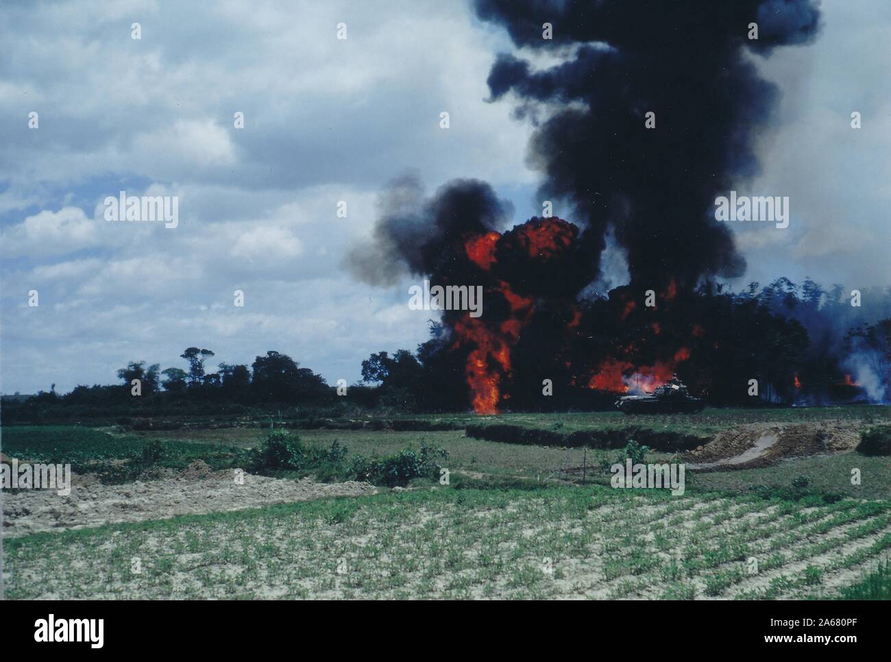 Ampio colpo, attraverso i campi verdi aperti, di un serviceman che guarda fuori dal tetto di un veicolo militare a un fiammeggiante, fumo esplosione al bordo di una zona boschiva, Vietnam, 1965. () Foto Stock