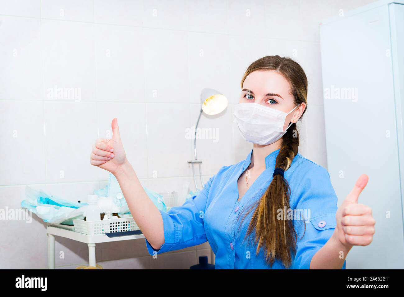 Attraente medico donna in maschera donna mostra Thumbs up segno a mano con sfondo ospedale Foto Stock