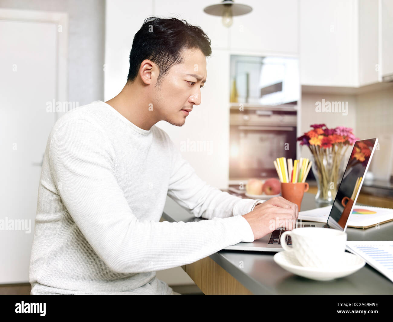 Giovane uomo asiatico che lavorano da casa seduti al bancone cucina utilizzando il computer portatile Foto Stock