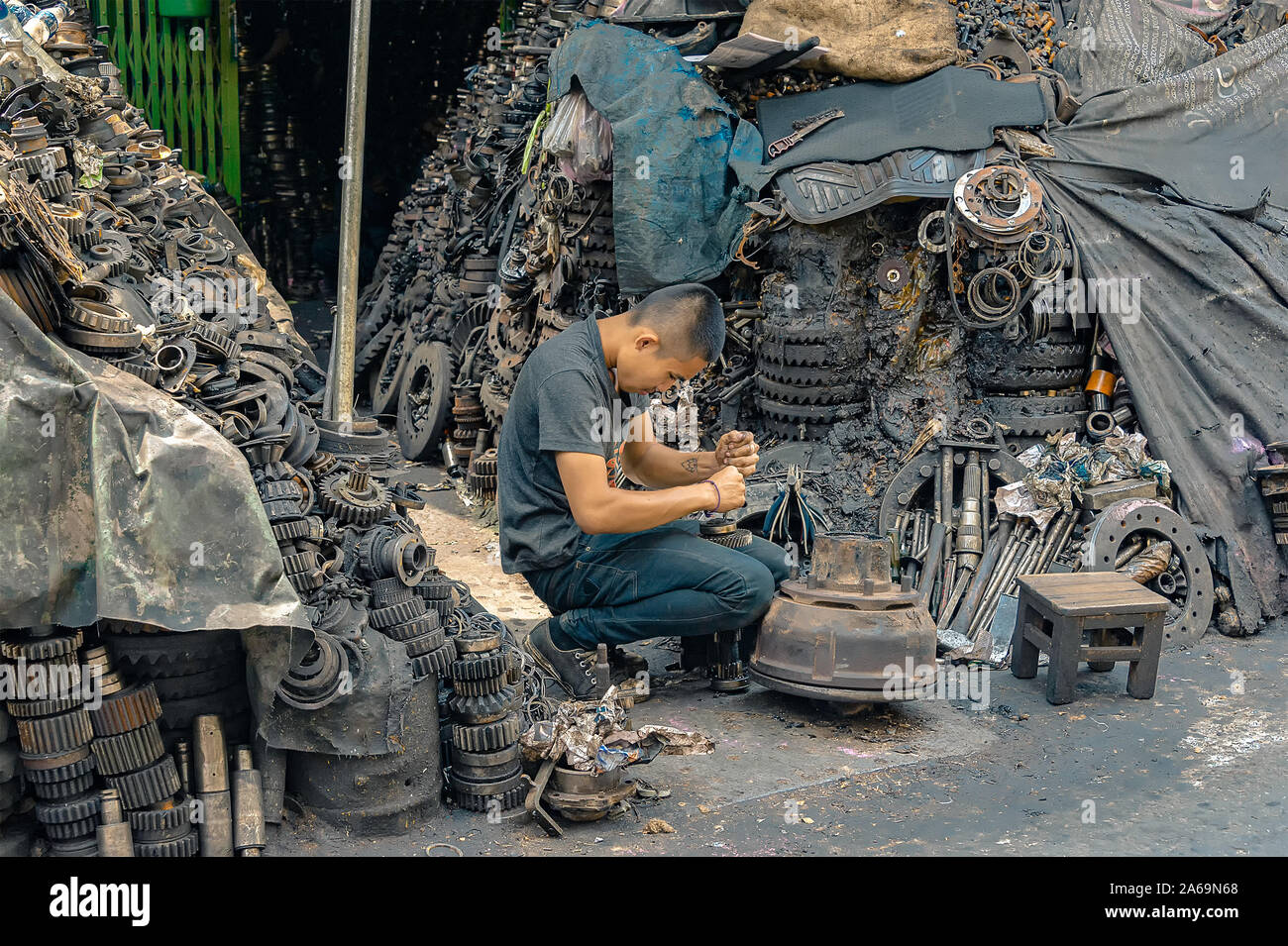 Bangkok - 28 Settembre 2019: un lavoratore smantella un motore in pezzi di vendere come pezzi di ricambio con la montagna dei pezzi del motore che lo circonda. Foto Stock