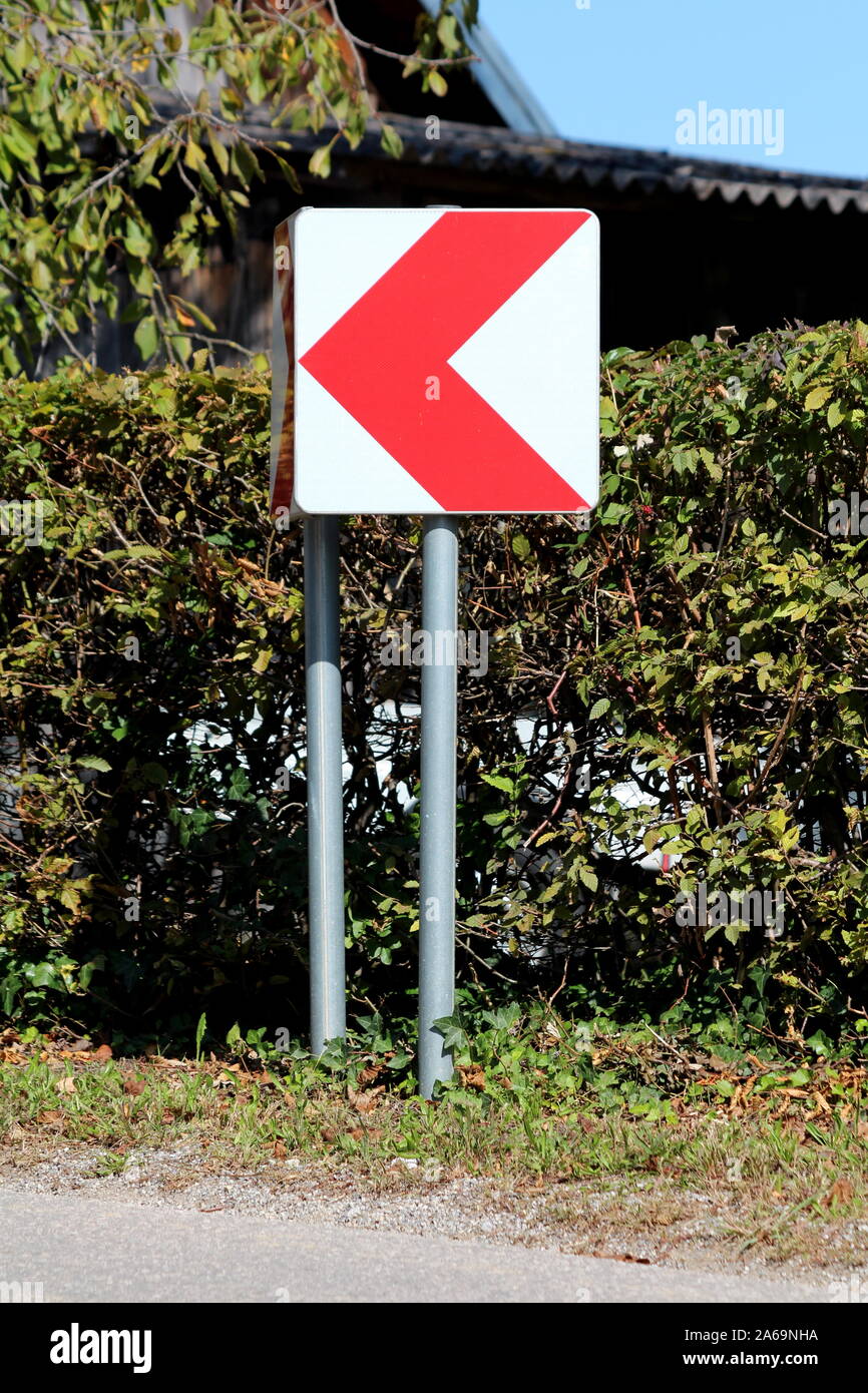 Curva pericolosa di colore rosso e bianco freccia cartello stradale montata su palo metallico in prossimità di strada pavimentata con siepe verde e casa in background Foto Stock