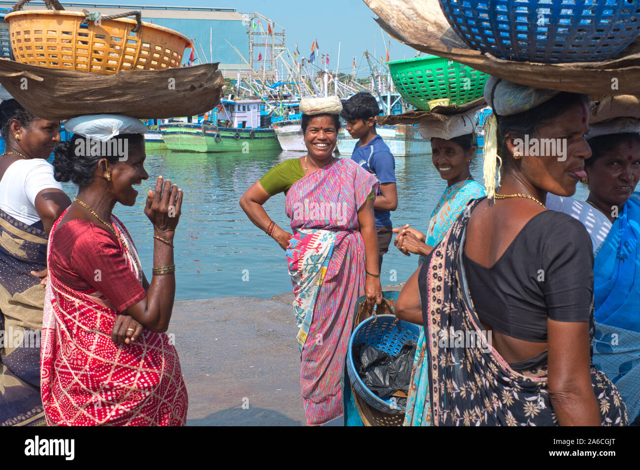 I venditori di pesci femmine che acquistano pesce al vecchio porto di Mangalore, Karnataka, India, si levano in piedi intorno scherzando, con le barche di pesca nello sfondo Foto Stock