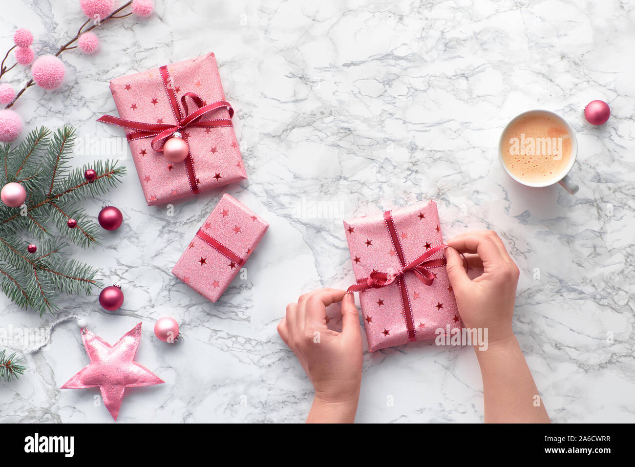 Piatto di Natale gettare sul tavolo di marmo. Legare le mani fino a nastro avvolto sul regalo di Natale. Tazza di caffè nelle vicinanze. Decorazioni invernali: Abete rami, rosa ninnoli e star Foto Stock