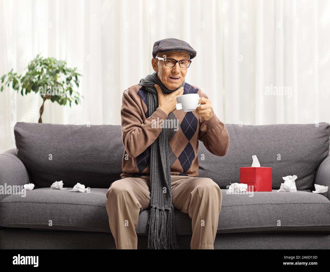 Senior con un influenza seduto su un divano a bere il tè e tenendo la sua gola Foto Stock