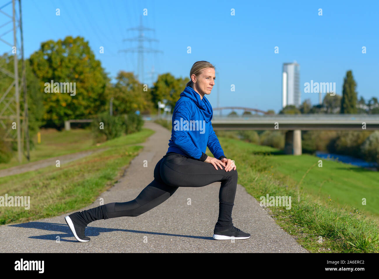 Montare athletic donna limbering fino a fare esercizi di allungamento per riscaldare i suoi muscoli su un percorso a fianco di un fiume in inverno in una giornata di sole Foto Stock