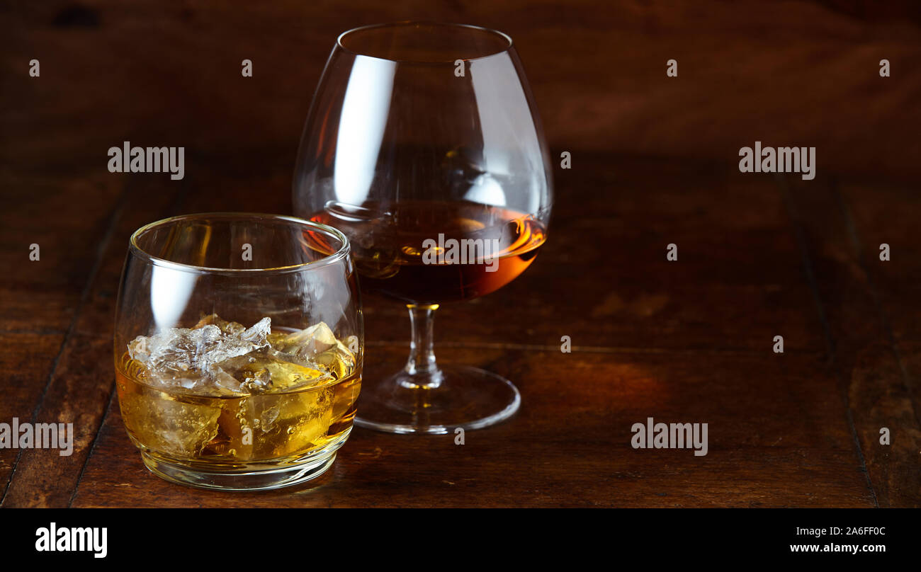 Whisky on the rocks e cognac in un snifter in piedi su una tavola di legno banco bar di notte con spazio di copia Foto Stock