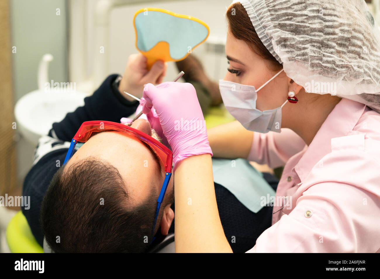 Dentista giovane donna considera un paziente un uomo. Il medico utilizza  guanti monouso e una maschera, cappello e occhiali di sicurezza. Il  paziente guarda nello specchio e che Foto stock - Alamy
