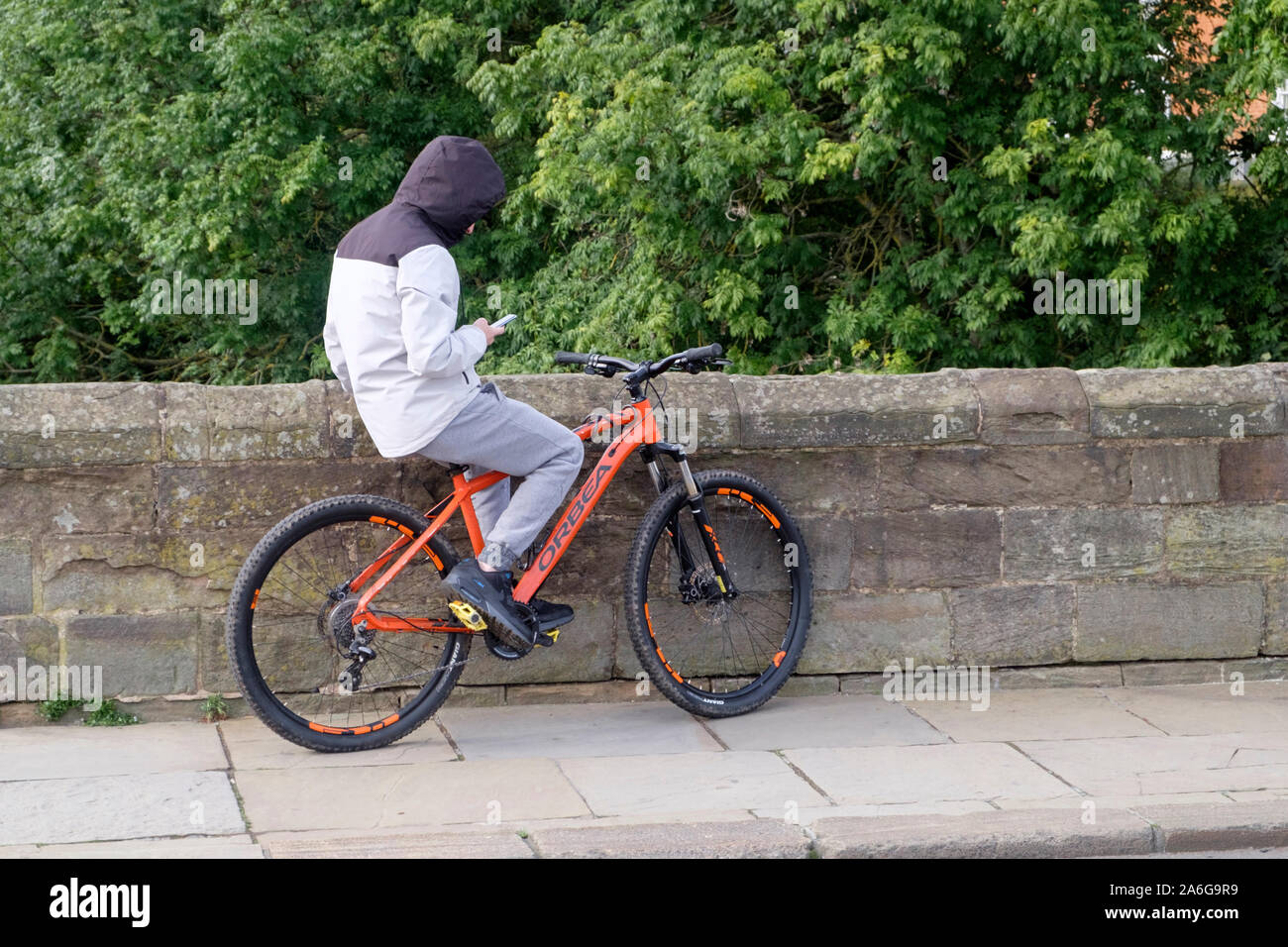 Giovane uomo o la gioventù di indossare una felpa con cappuccio sat sulla sua bicicletta guardando al suo telefono cellulare Foto Stock
