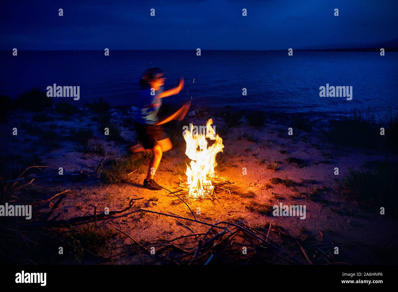 Giovane uomo è il salto al di sopra del campo di fuoco sulla spiaggia vicino al lago al crepuscolo Foto Stock