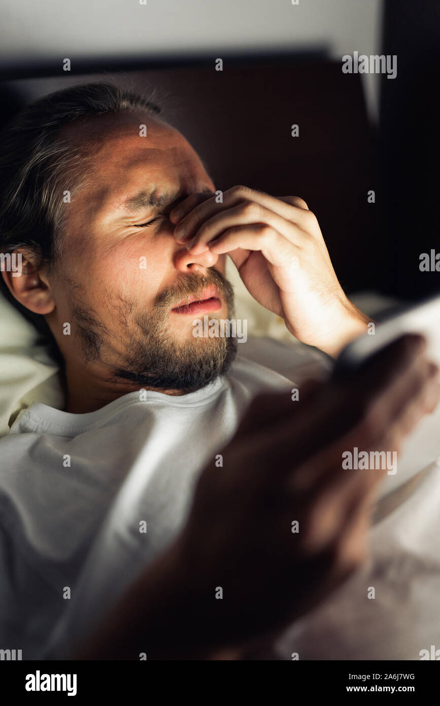 Barbuto maschio caucasico è guardare qualcosa sul suo tablet a letto a notte. Egli è stanco, non riesce a dormire e lui ha chiaramente un mal di testa. Foto Stock