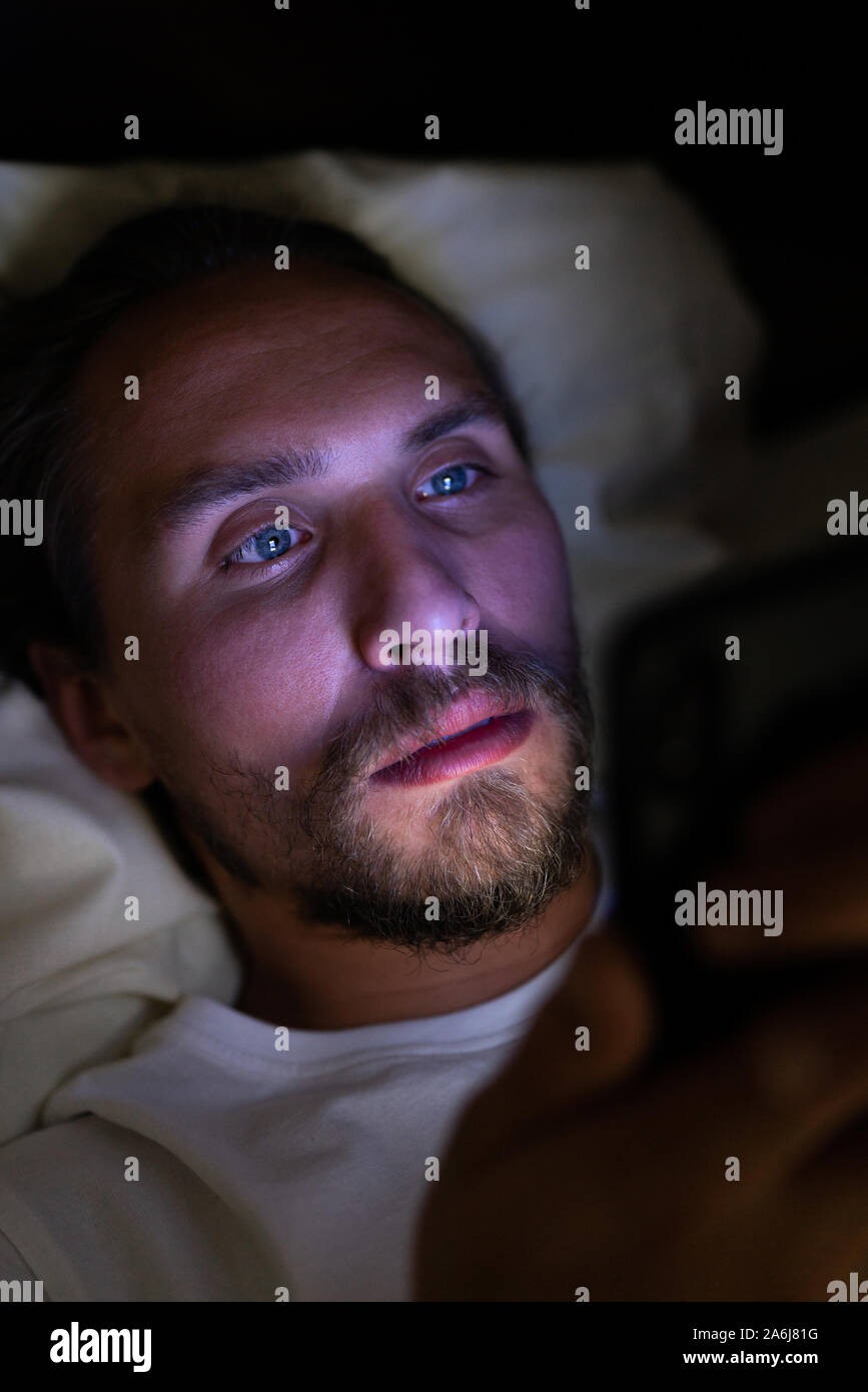 Giovani belli e stanchi con barba non riesce a dormire e guardare qualcosa sul suo telefono cellulare di notte. Foto Stock
