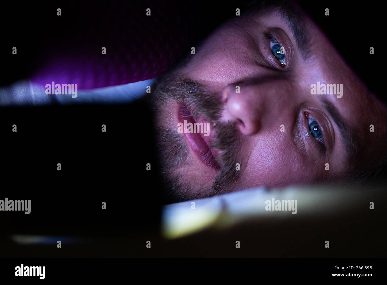 Barbuto giovane uomo è disteso nel letto sotto la sua coperta. Egli non riesce a dormire e sta guardando qualcosa sul suo telefono cellulare. Foto Stock