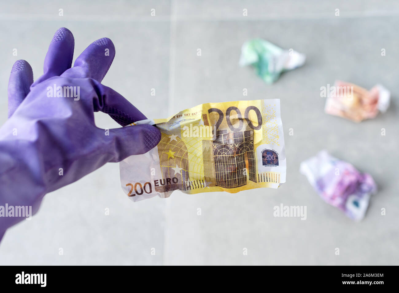 La mano in viola i guanti di gomma per la pulizia azienda accartocciata euro bills su sfondo grigio. Pagamento per lavoro, cestino di denaro, l'inflazione, il tema dei rifiuti. Foto Stock