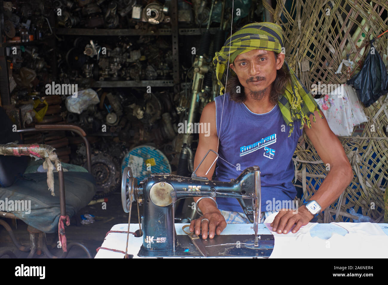 Un sarto con la sua vecchia macchina da cucire a pedale che occupa un piccolo spazio di fronte ad un negozio di rottami di metallo; Bhendi Bazar, Mumbai, India Foto Stock