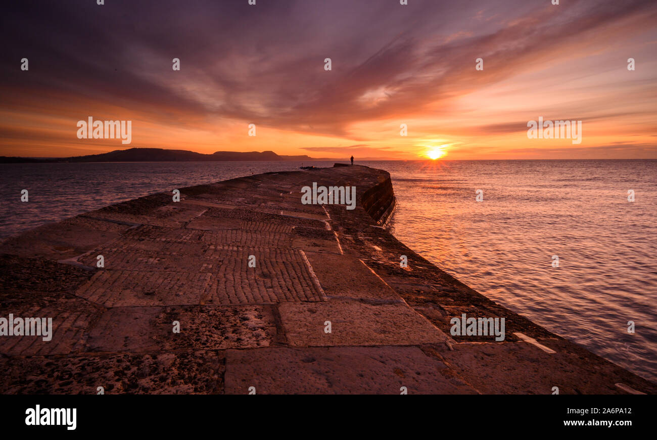 Lyme Regis, Dorset, Regno Unito. 28 ott 2019. Regno Unito Meteo: bellissima alba presso il Cobb Lyme Regis. Un uomo guarda su come il cielo sopra la storica parete Cobb si illumina con colori brillanti su una fredda mattina autunnale. Credito: Celia McMahon/Alamy Live News Foto Stock