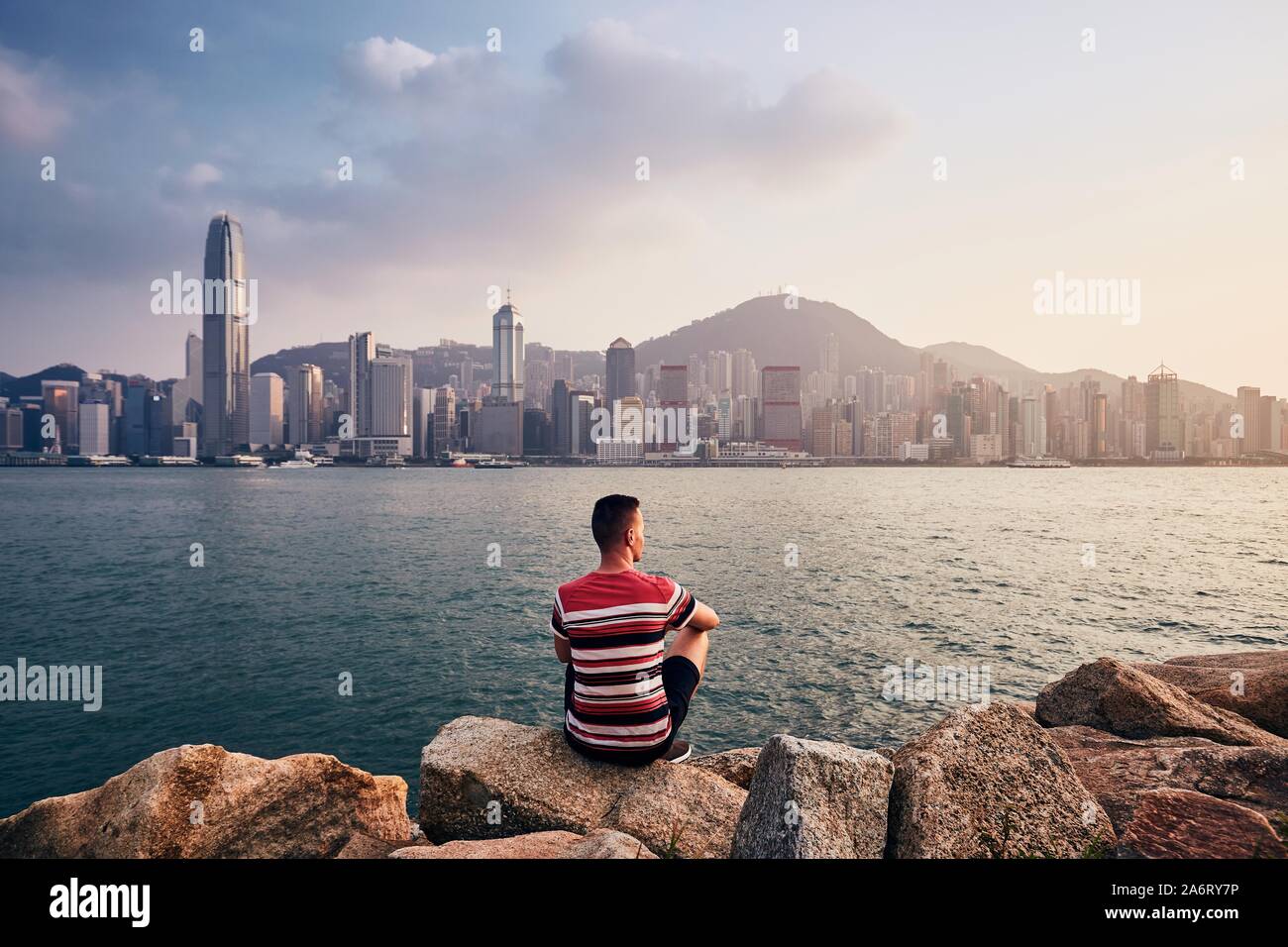 Giovane uomo seduto sulle pietre contro Hong Kong cityscape con grattacieli al tramonto colorato. Foto Stock