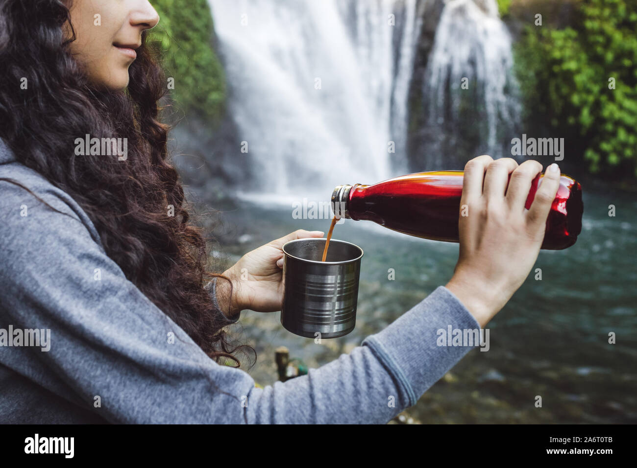 Donna mani versando il caffè caldo dal rosso thermos in acciaio tazza. Sfondo della cascata tropicale. Rifiuti Zero eco concetto. Attrezzatura per le escursioni. Foto Stock