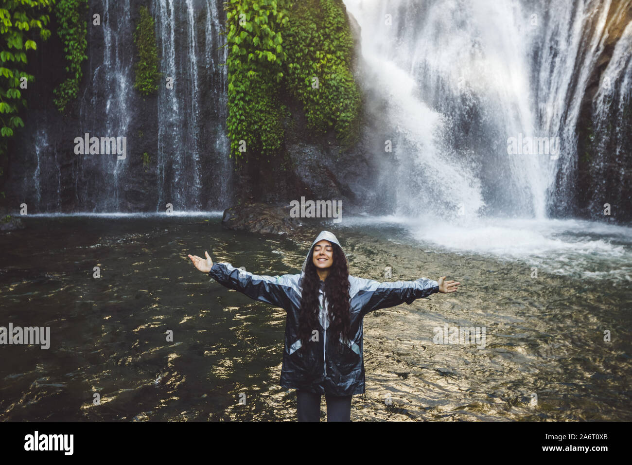 Giovane donna felice godendo di una cascata di Bali. Indossando un impermeabile grigio da spruzzi d'acqua. Viaggi in Indonesia. Foto Stock