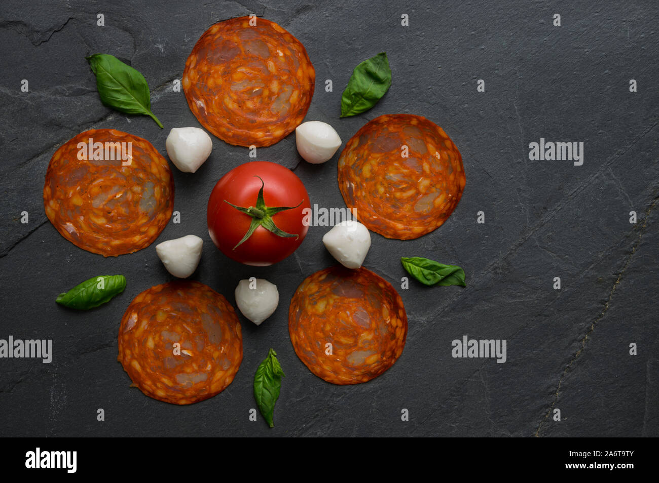 Tradizionale pizza italiana ingredienti mozzarella, salame, pomodoro e basilico foglie composte in una forma di fiore su nero pietra naturale dello sfondo. Foto Stock