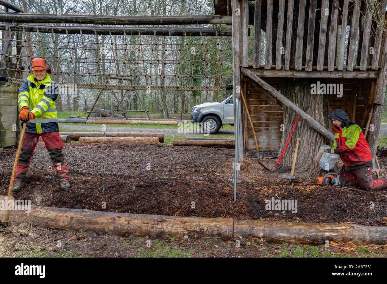 Registratori di giovani a lavorare con un'ascia su un parco giochi, tenendo un tronco di albero di taglio, mentre gli altri lavoratori riempie la benzina in una seghe a catena nel giorno di pioggia. Foto Stock