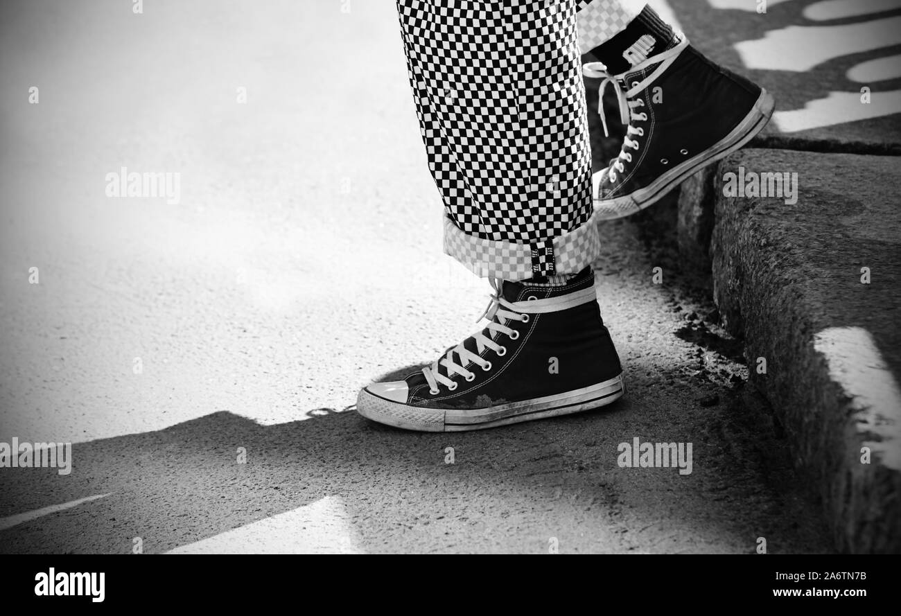 Un'immagine in bianco e nero dei piedi di un uomo discendente, vestito nel quartiere alla moda di plaid pantaloni e scarpe da ginnastica, illuminato dalla luce solare. Foto Stock