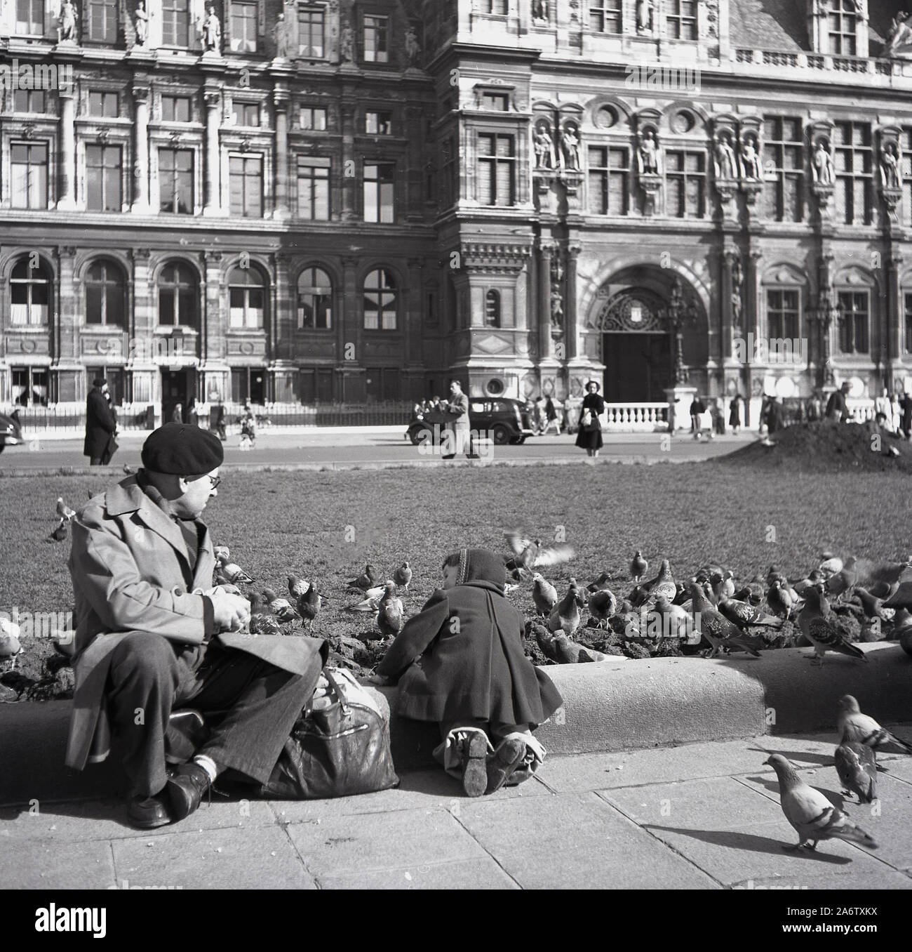 1950, storico, un gentiluomo parigino in un berretto e un impermeabile con il suo grande titolo seduto a dare da mangiare ai piccioni sulla piazza di fronte al Grand Hotel de Ville, il municipio di Parigi, Francia. Foto Stock