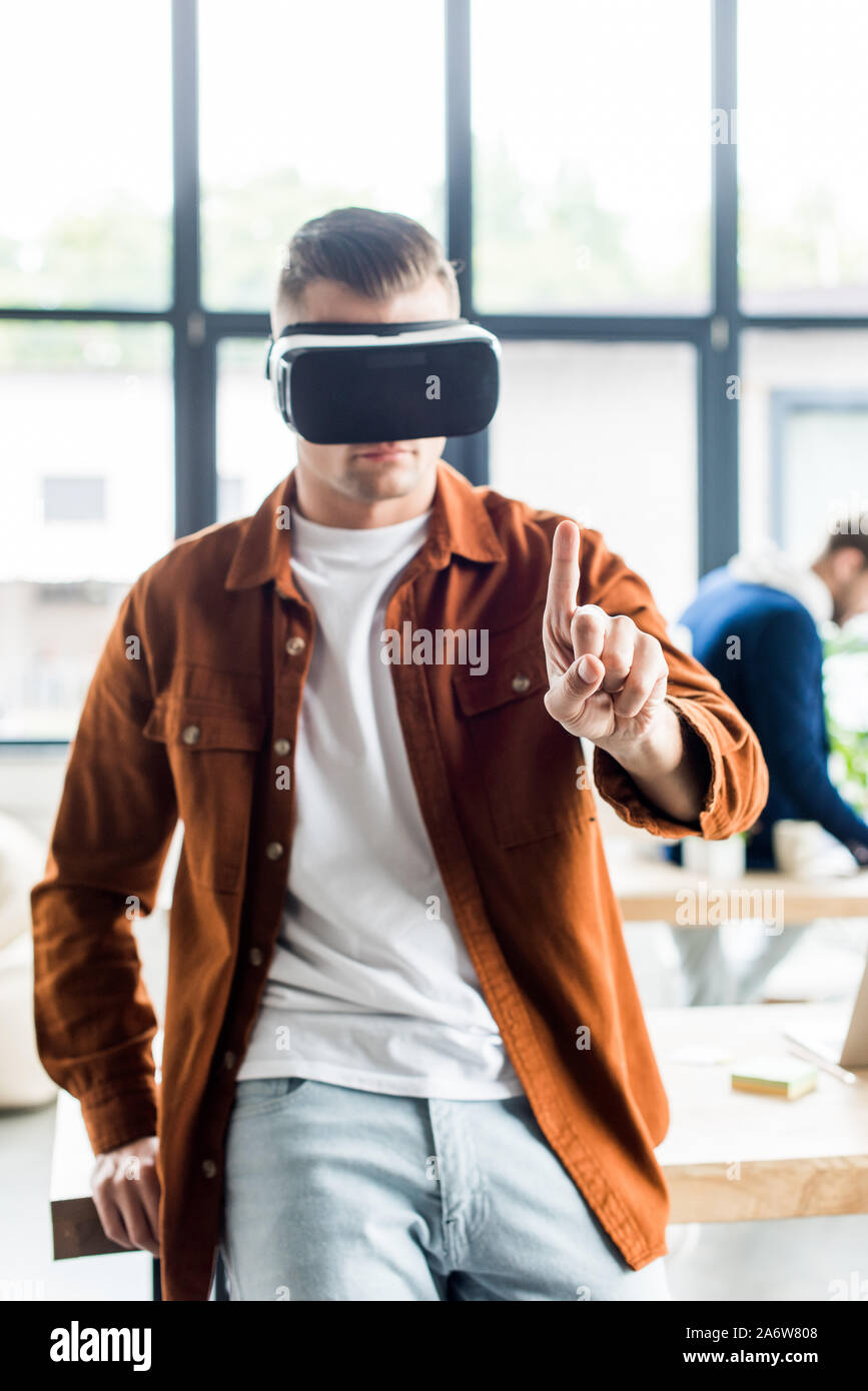 Giovane imprenditore di toccare qualcosa con il dito mentre utilizzando la realtà virtuale auricolare in carica Foto Stock