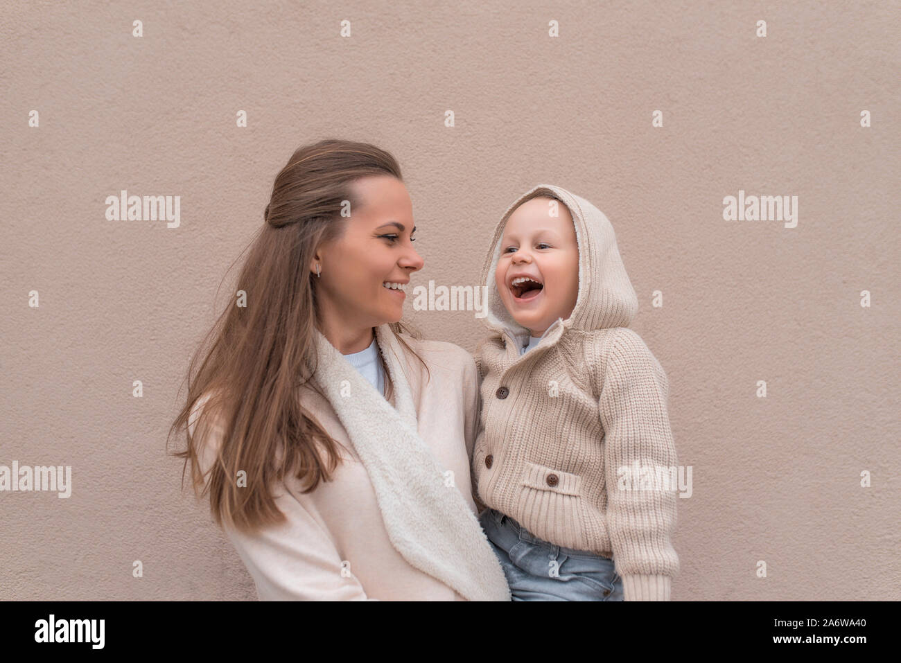 La famiglia felice la mamma donna con little boy, ridendo, sorridente, divertendosi, la riproduzione. Sfondo muro beige. Caldo abbigliamento casual, felpa con cappuccio. Spazio libero Foto Stock