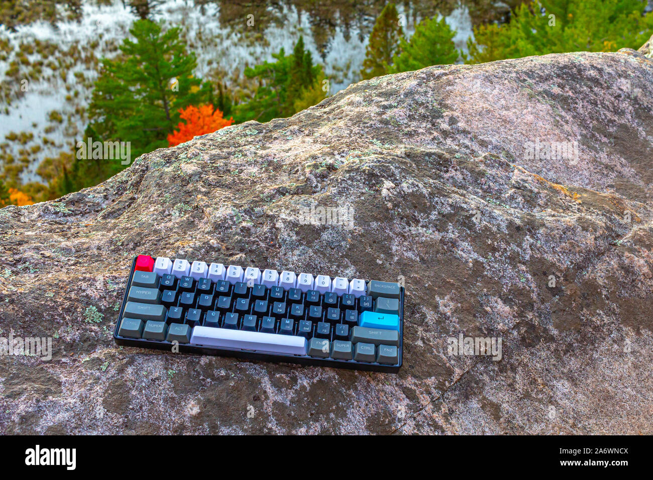 Una meccanica della tastiera del computer con il grigio, bianco, nero, rosso e tasti blu sembra un po' fuori posto sulla roccia di una rupe sopra gli alberi e un laghetto in Foto Stock