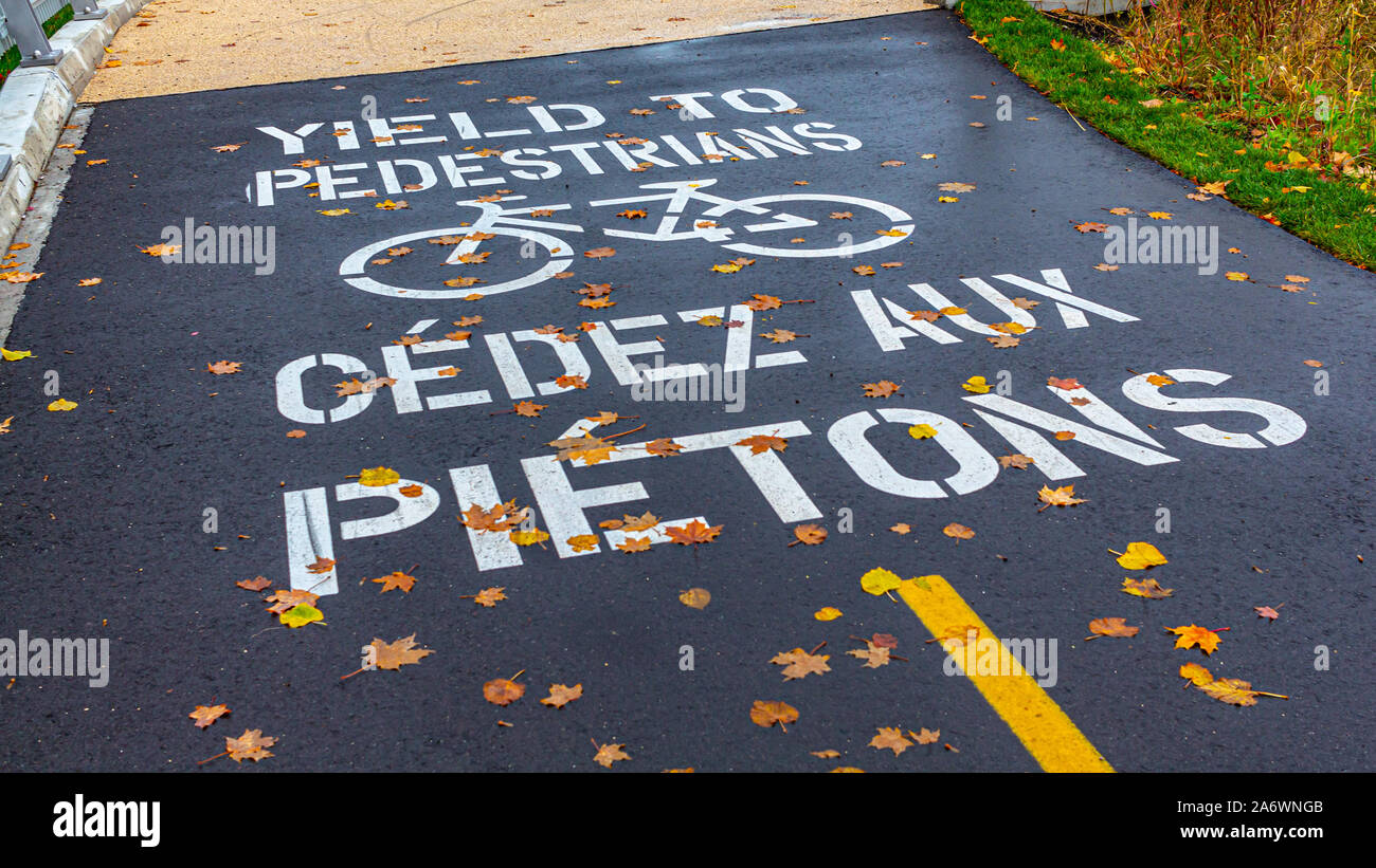 Un avvertimento stampata su un asfalto percorso avverte le biciclette per dare ai pedoni a piedi sia in inglese che in francese. Foto Stock