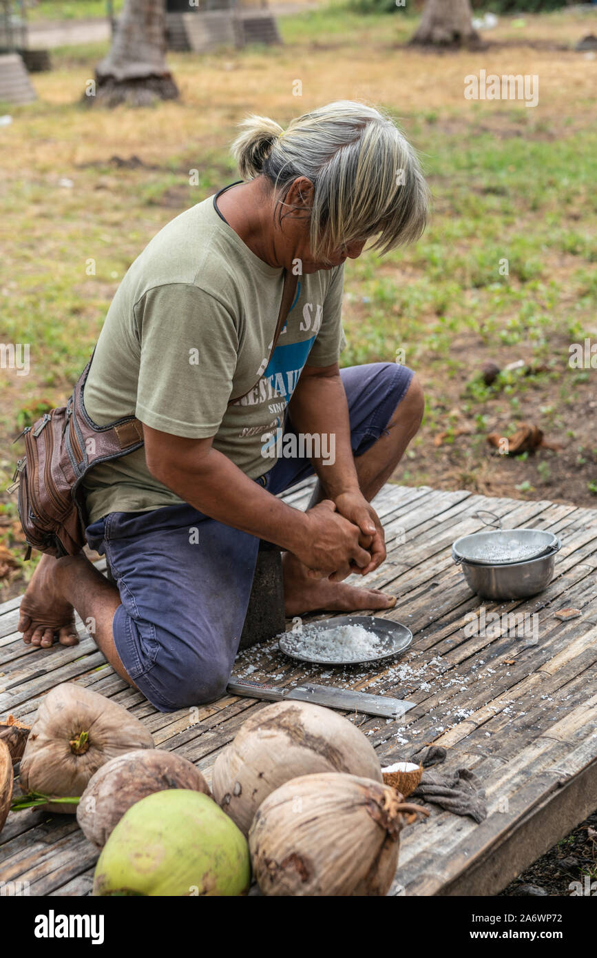 Ko Samui, Tailandia - 18 Marzo 2019: dai capelli grigi uomo esegue la fase quattro della elaborazione di cocco: carne di raschiatura, copra, fuori della metà utilizzando bla Foto Stock