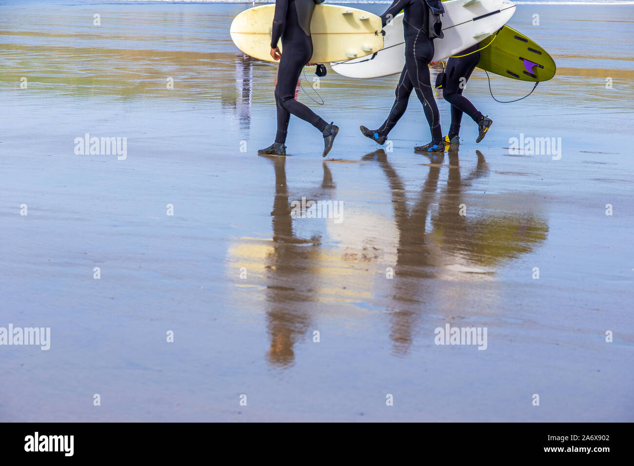 Surfers, trasportando i surfboards, camminando lungo una spiaggia bagnata a Polzeath, Cornovaglia, Regno Unito Foto Stock