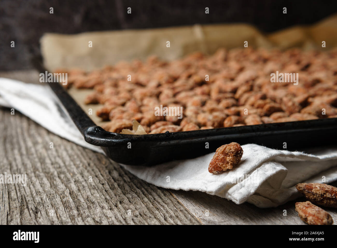 Mandorle bruciato esteso su nero vassoio da forno, carta, tessuto bianco sulla trama rustico tavolo in legno, concentrarsi su tre bruciate le mandorle in primo piano, scuro Foto Stock