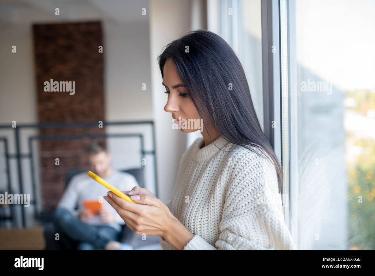 Dai capelli scuri moglie in piedi vicino alla finestra utilizzando il telefono Foto Stock