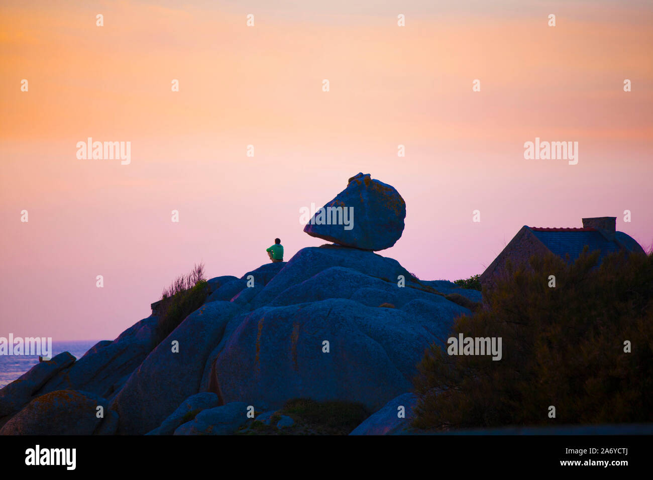 Ein junger Mann beobachtet den Sonnenuntergang am Pointe de Pontusval in der Bretagne. Neben ihm ein tonnenschwerer, riesiger Felsbrocken, der wirkt, Foto Stock