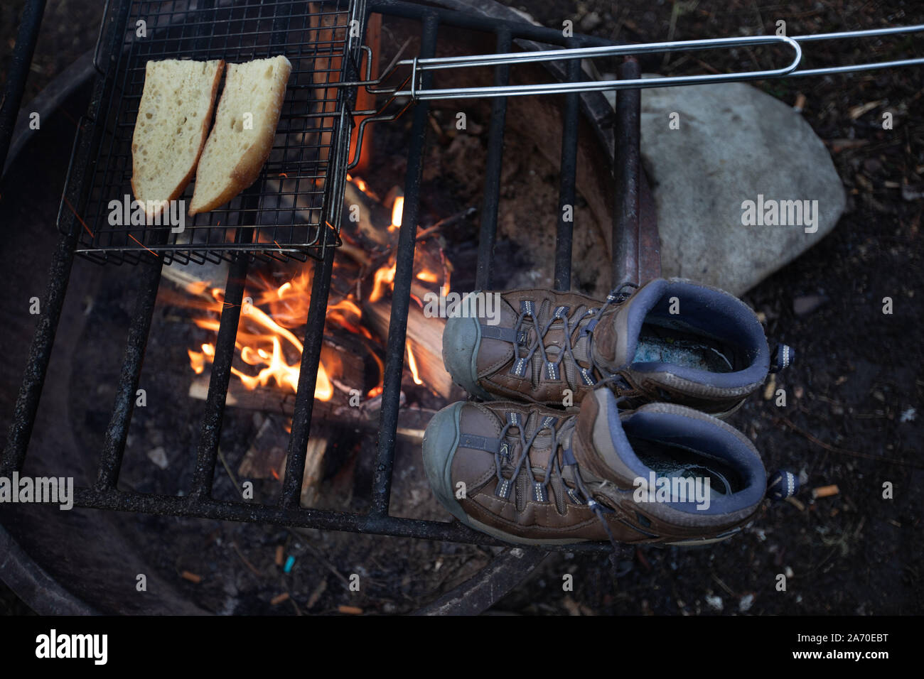 Wet scarpe da escursionismo essiccamento mediante un falò con la tostatura del pane sulle fiamme. Foto Stock