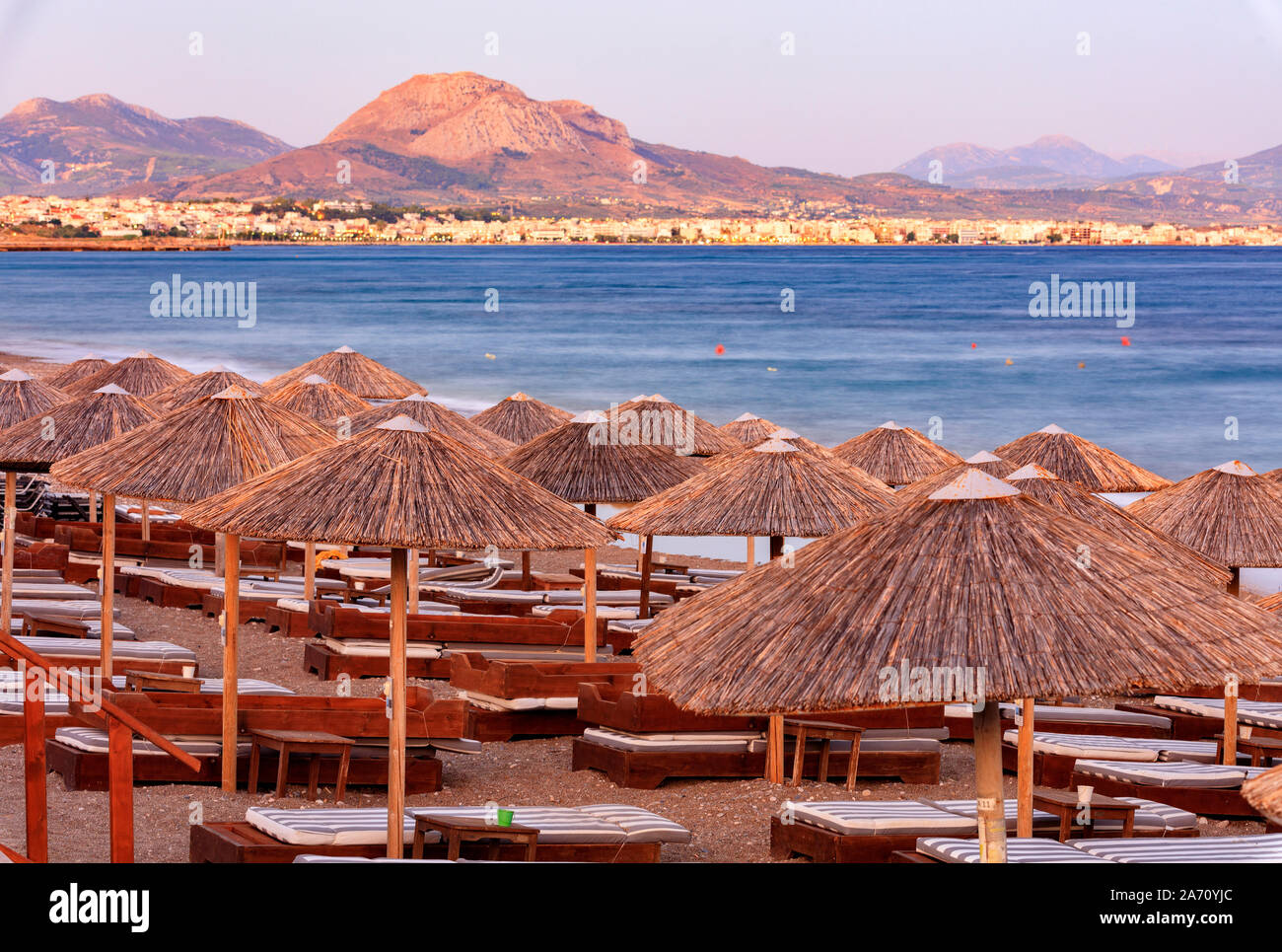 I tetti di paglia di ombrelloni in legno e sedie a sdraio con materassi su una spiaggia deserta del lungomare di raggi del tramonto. Foto Stock
