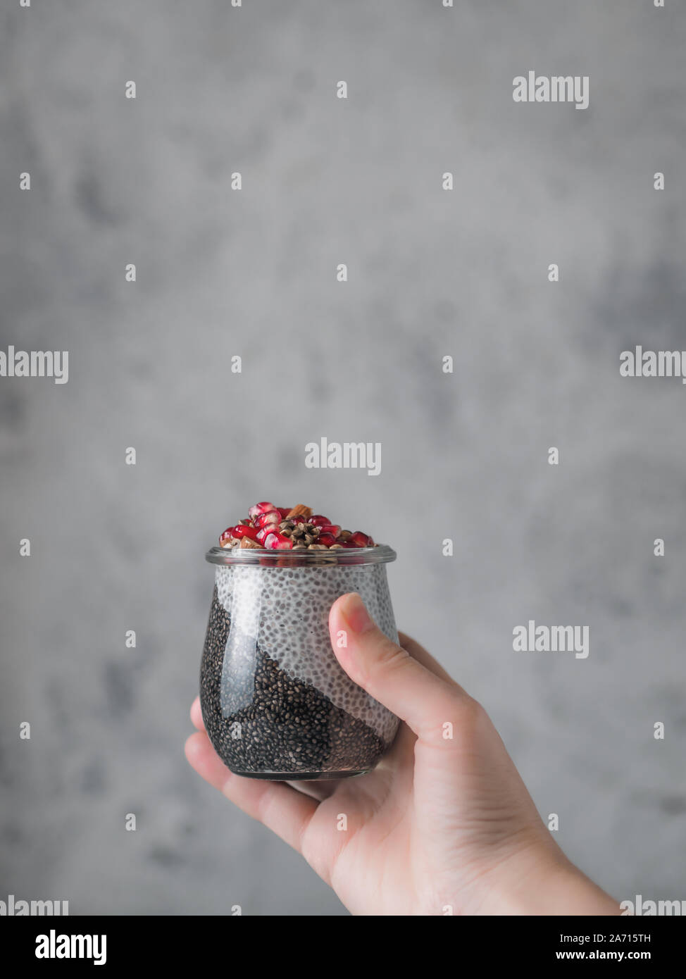 Budino di Chia con il nero di carbone bianco e vaniglia a base di vegetali in latte di donna mano su sfondo grigio. Femmina di tenere in mano il vasetto di vetro con chia puding servita melograno e la canapa. Copia dello spazio. In verticale Foto Stock